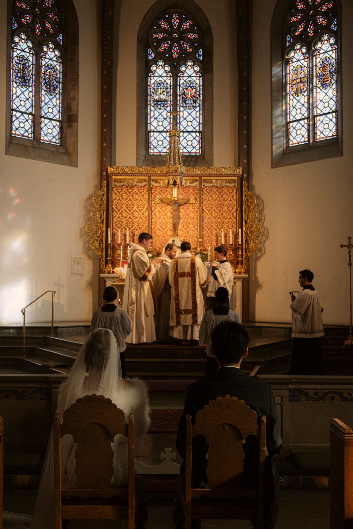 Bride and groom kneel before priests at ornate golden altar with stained glass windows during Mass.