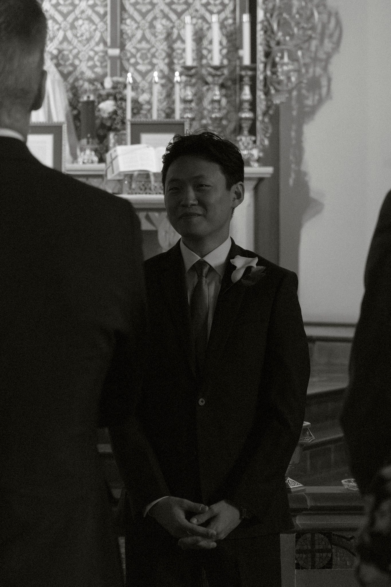 Black and white photo of a young man in a suit with boutonniere smiling at a church wedding ceremony.