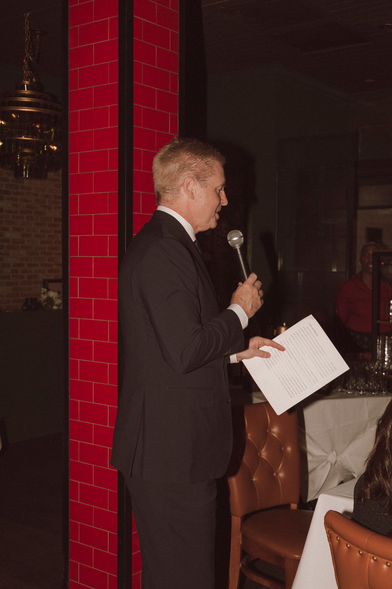 Blonde man in black suit delivering a speech at a wedding reception while holding a microphone and papers.