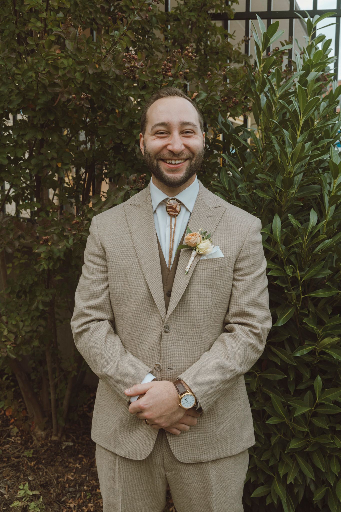 A groom in a tan suit poses against a backdrop of green shrubs.