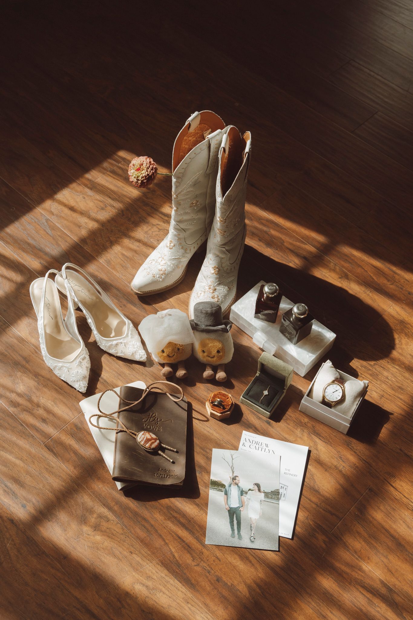 Wedding accessories including white cowboy boots, heels, jewelry, and photographs arranged on a wooden floor in sunlight.