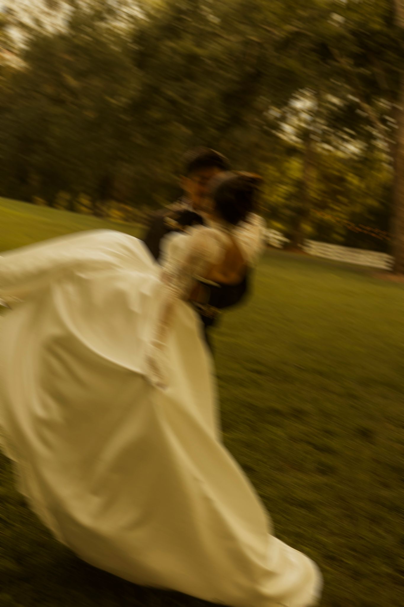 Blurred motion photo of bride in white gown and groom dancing joyfully on green grass at outdoor wedding.