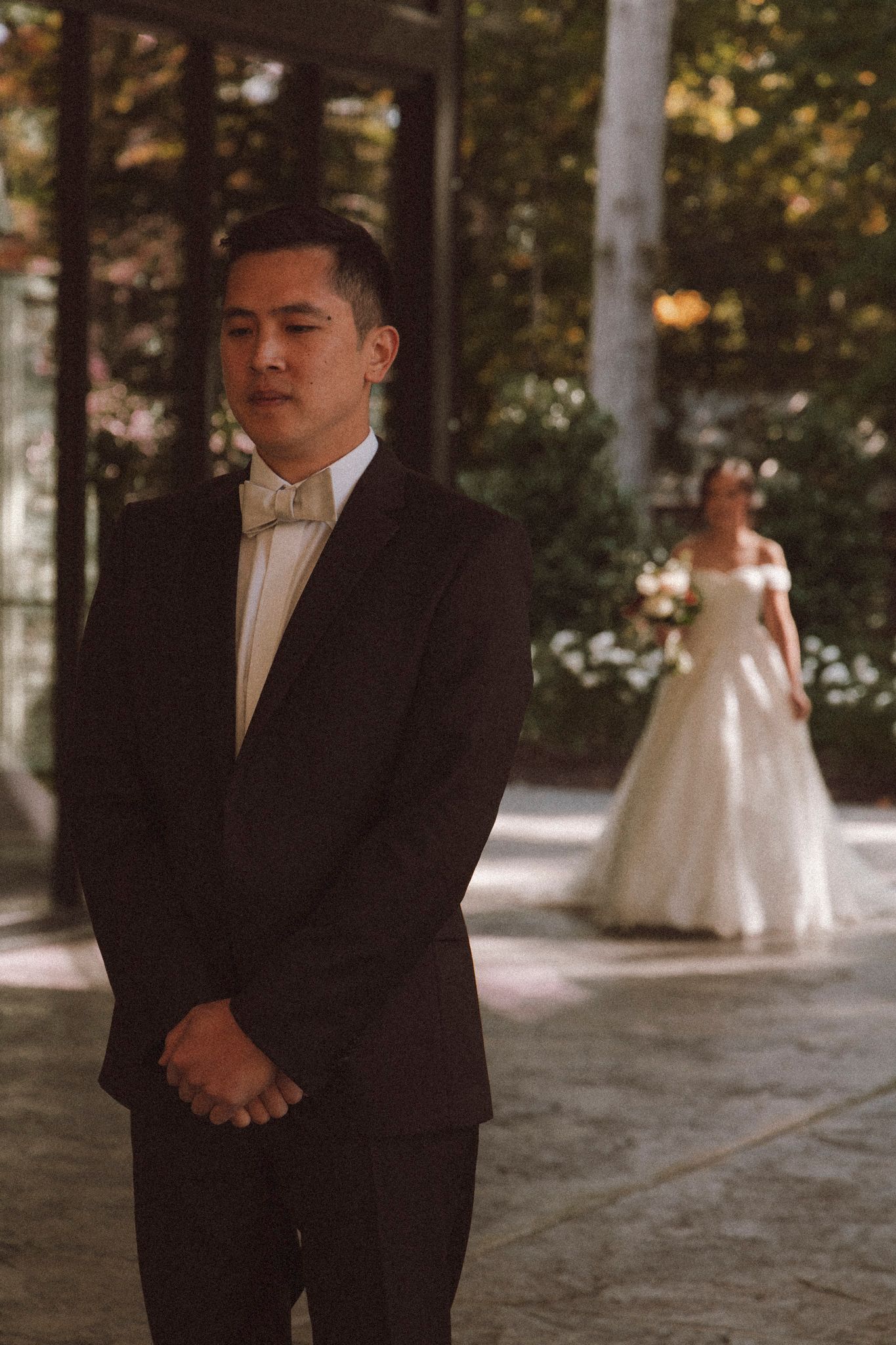 A person in a black suit stands in a formal setting with a bride in a white dress visible in the background.