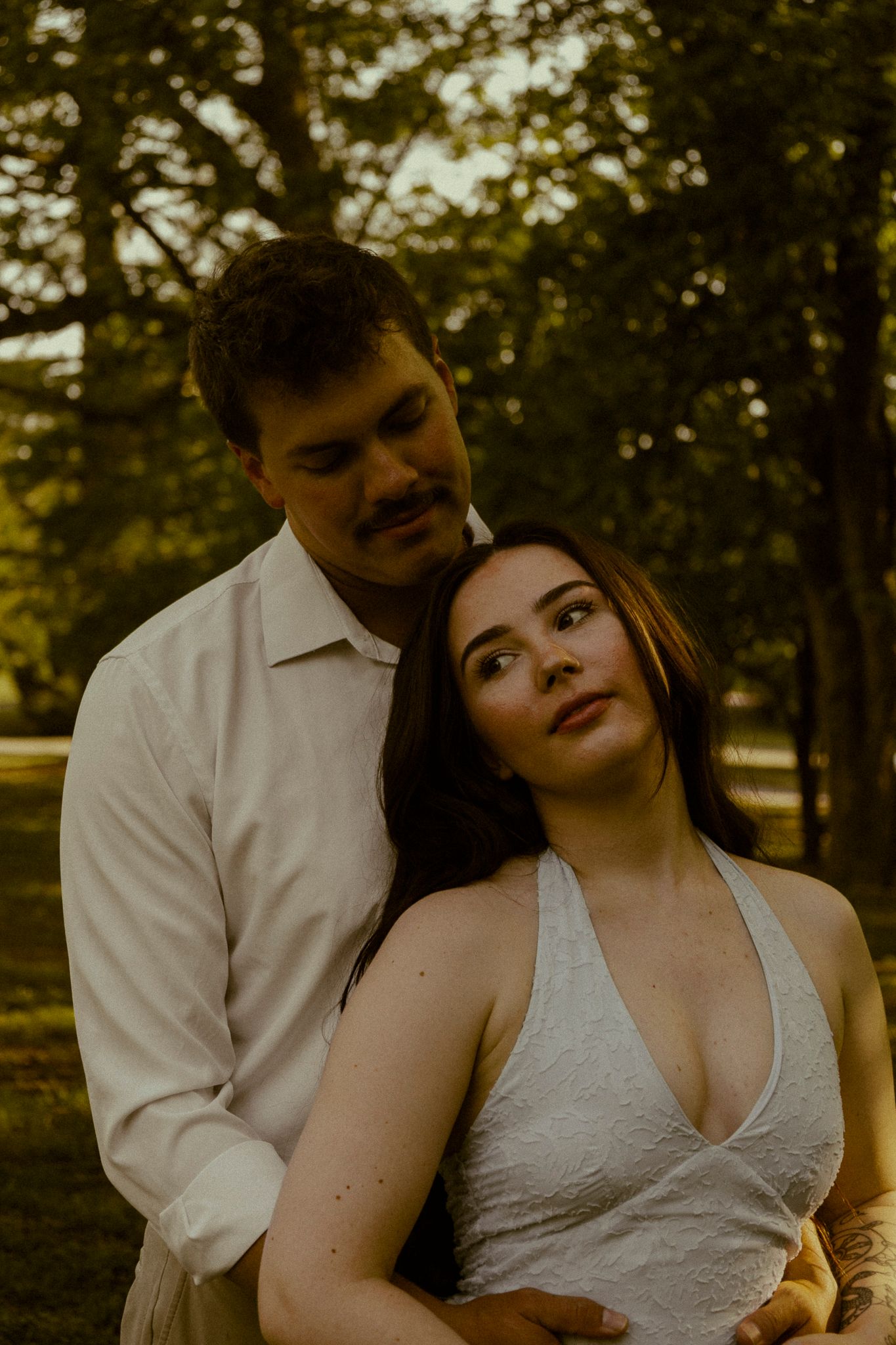 Romantic couple embracing outdoors in a park, woman in white halter dress leaning back against man in white shirt.