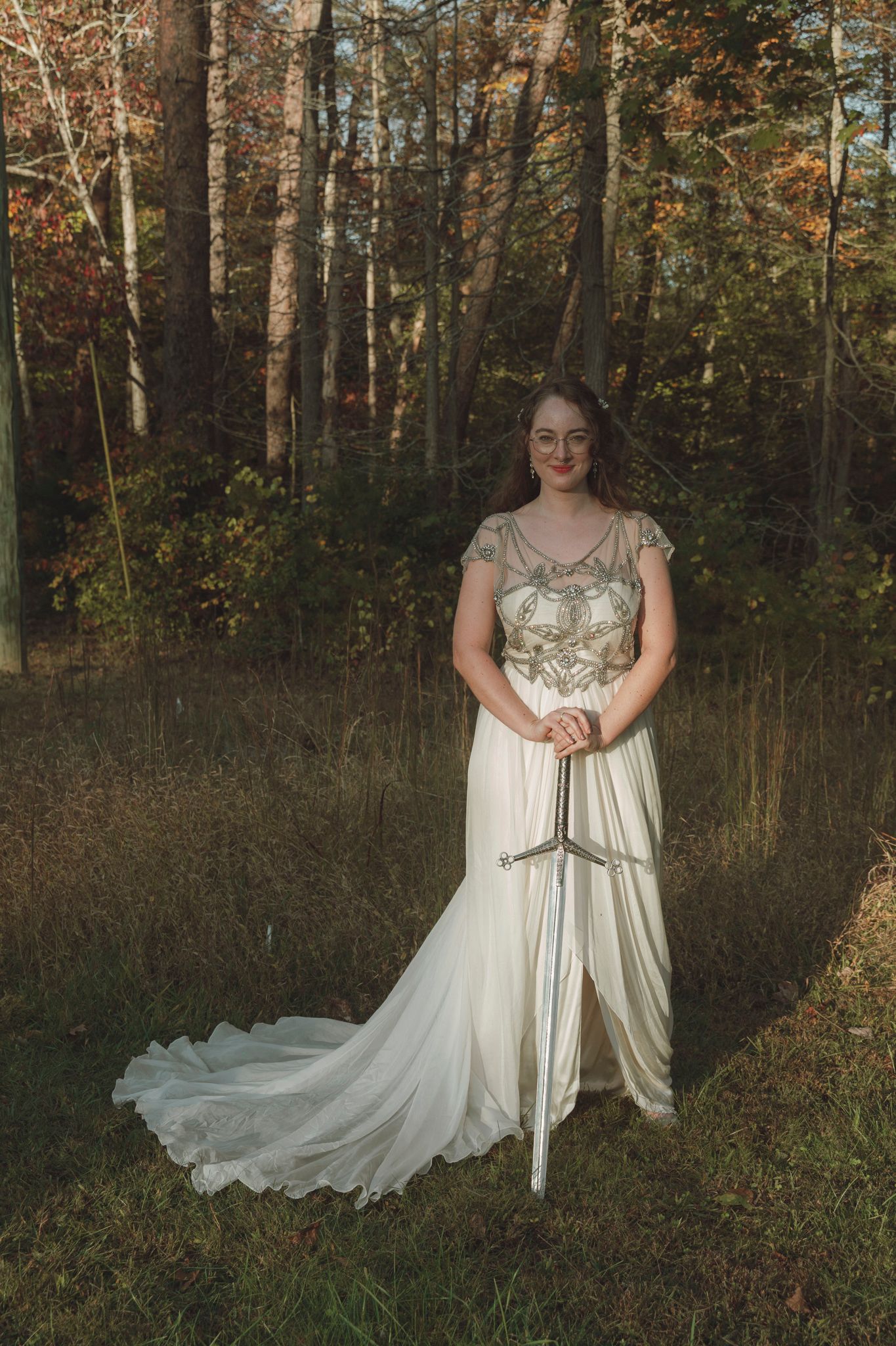 A person in a flowing white wedding dress with a train stands in a sunlit autumn forest holding a sword.
