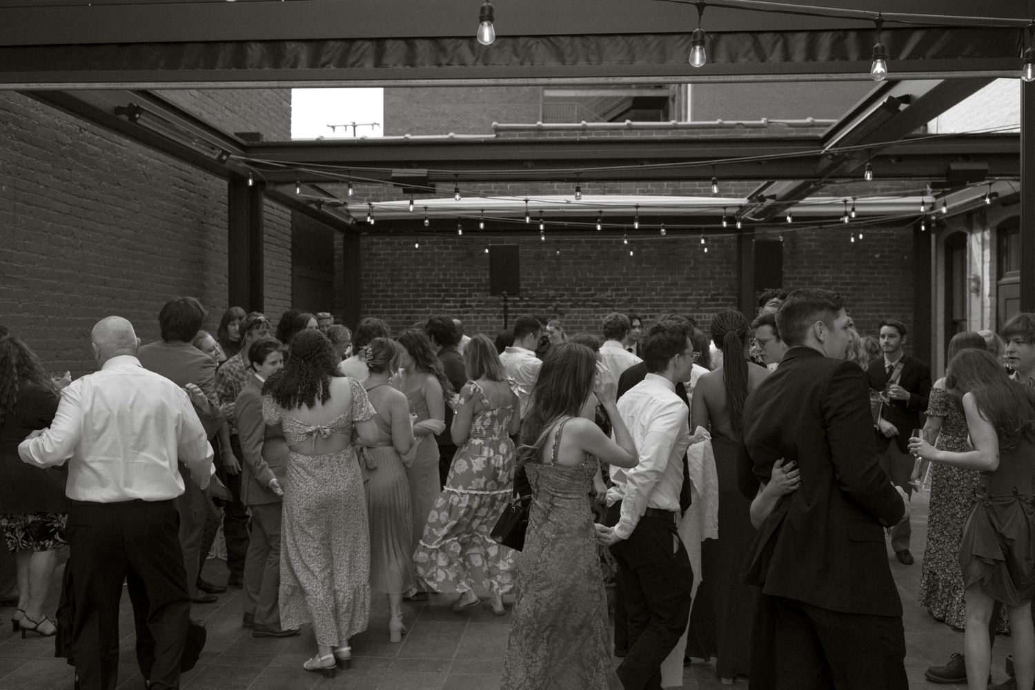 A black and white photo shows people dancing and mingling at an indoor event under industrial ceiling beams.
