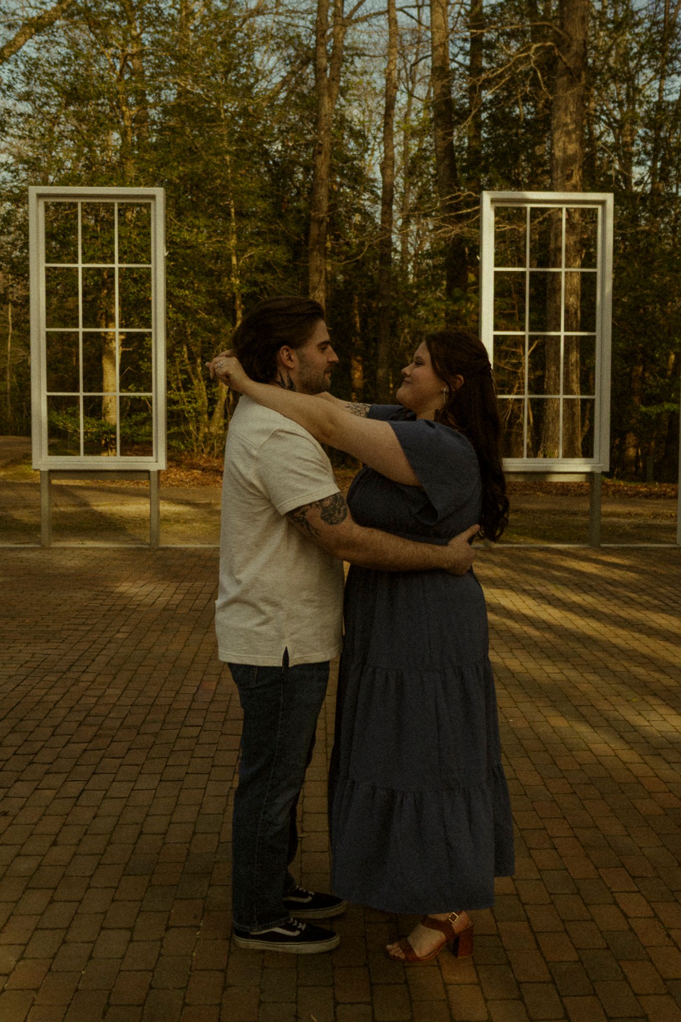 Couple embracing outdoors on brick path with white window frames and tall trees in background at golden hour.