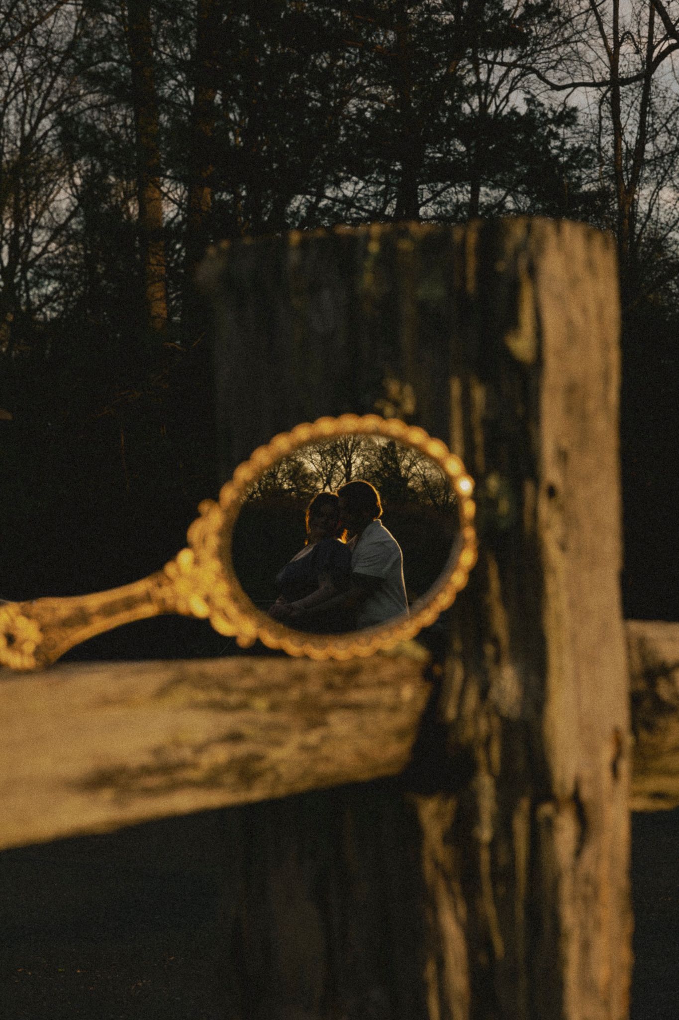 Romantic couple reflected in a vintage gold hand mirror resting on a rustic wooden fence at dusk.