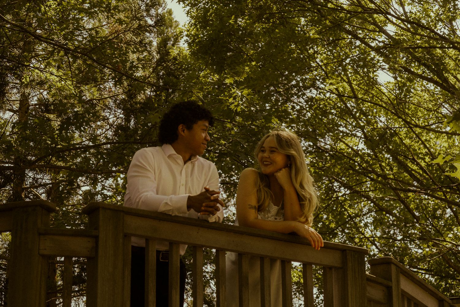Couple smiling and leaning on wooden deck railing surrounded by lush green trees in a romantic outdoor setting.
