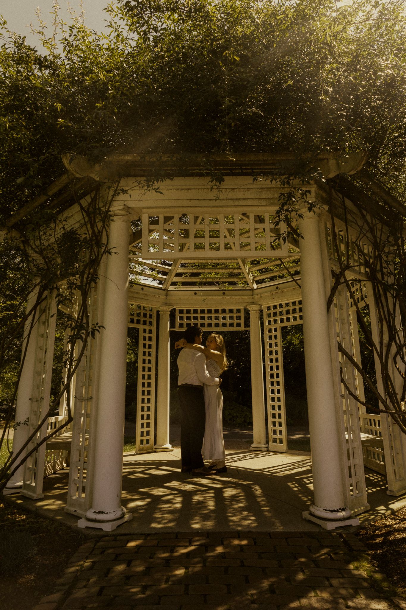 Couple embracing under a white lattice garden gazebo covered in lush green vines in warm golden sunlight.