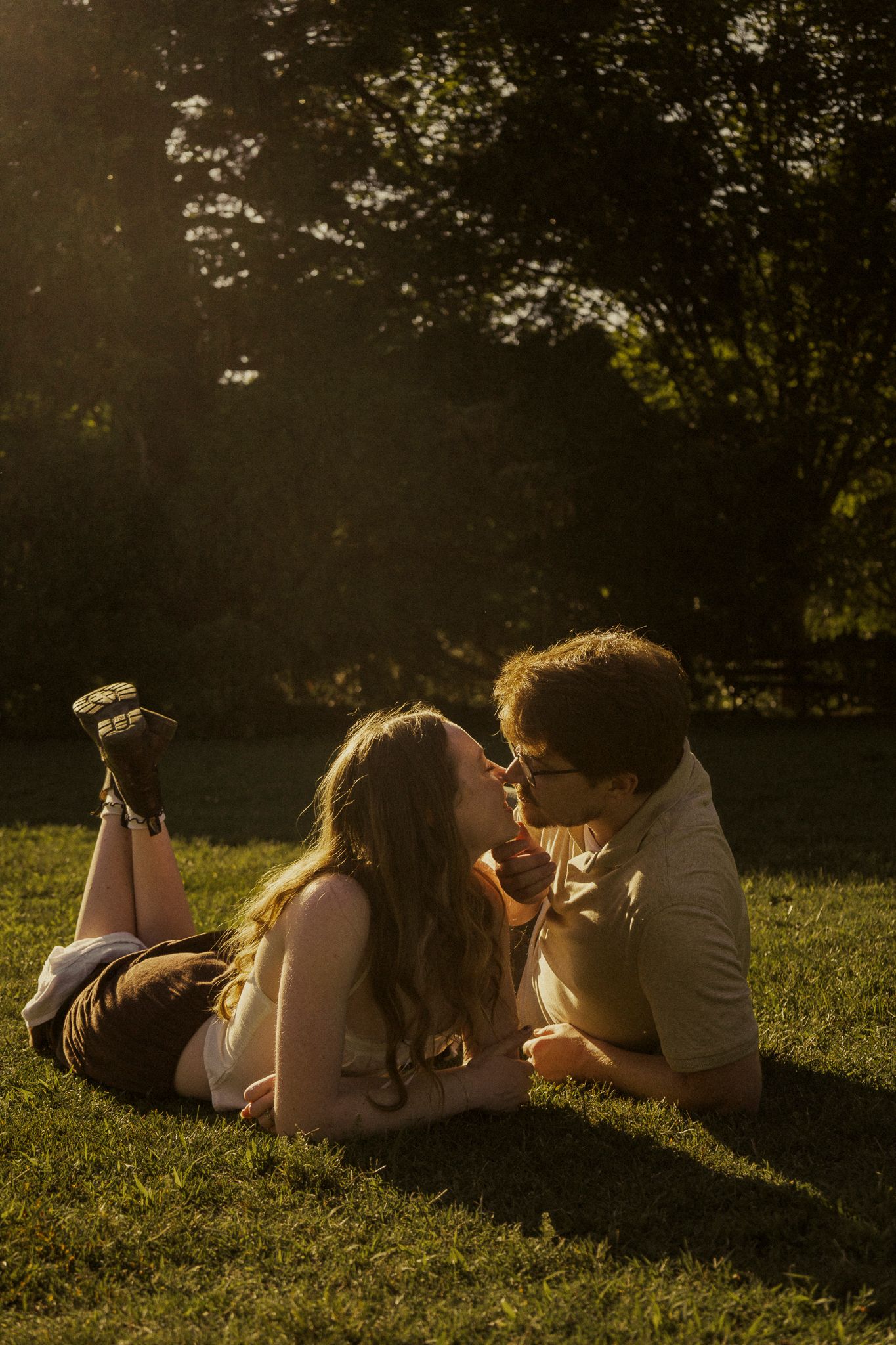 Couple sharing a romantic kiss while lying on green grass in golden hour sunlight with trees in background.