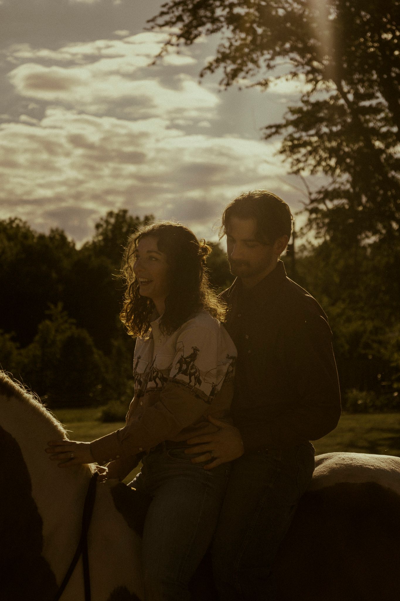 Couple riding a white horse together at golden hour sunset with trees and dramatic clouds in background.