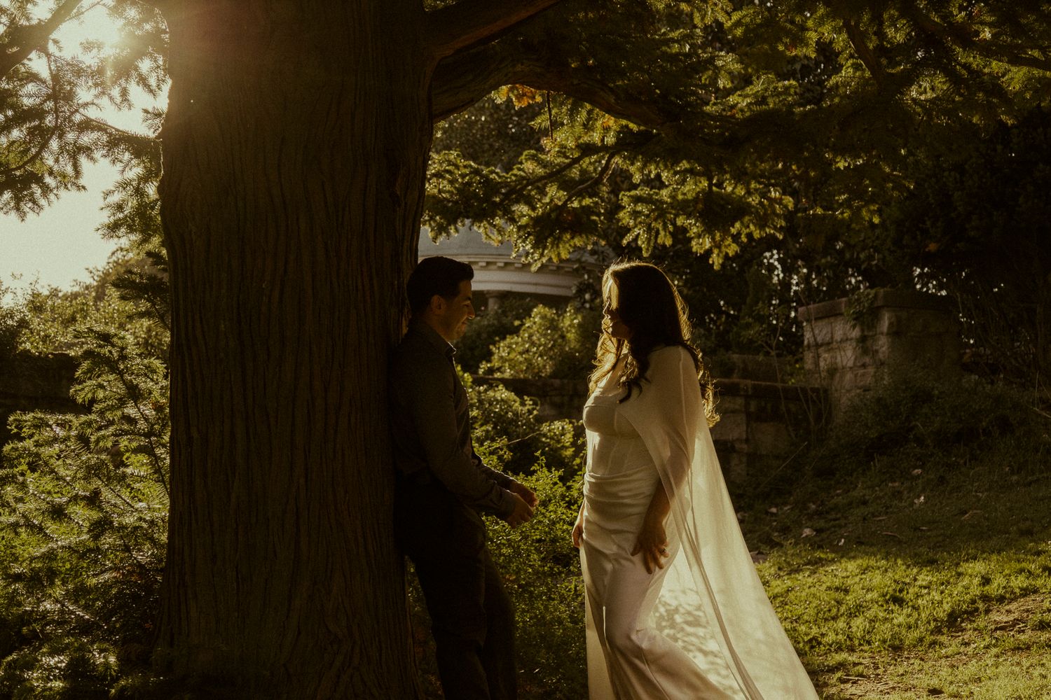 Couple sharing a romantic moment under a large tree in golden sunset light, bride in white gown.