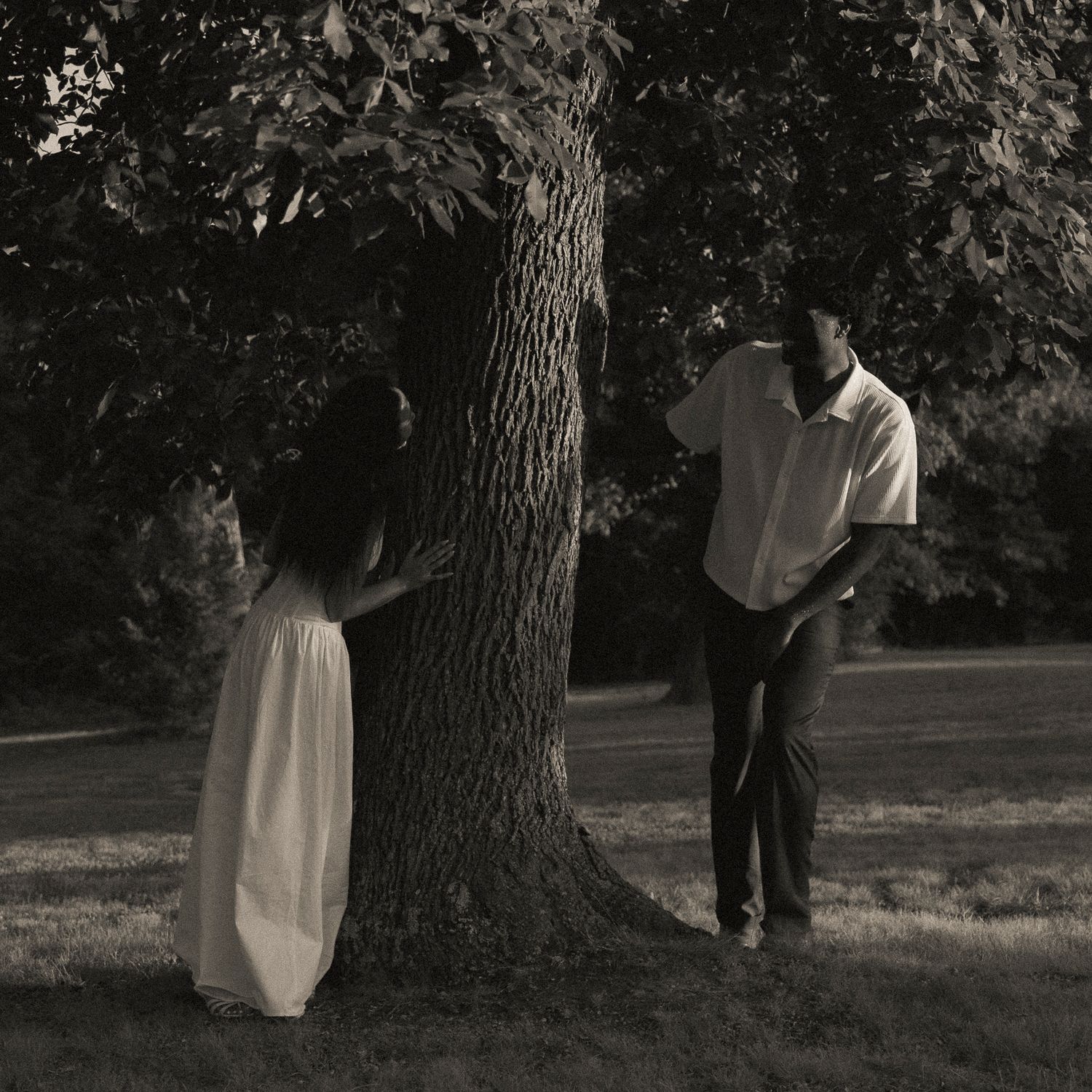 A couple stands on opposite sides of a large tree in a park, captured in a romantic sepia-toned photograph.