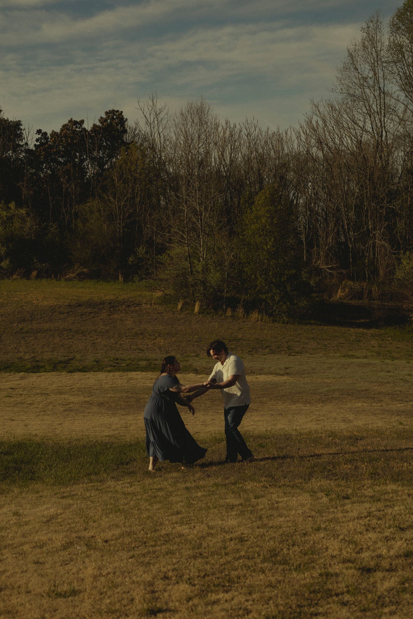 Couple dancing playfully in a sunlit autumn field surrounded by bare trees.