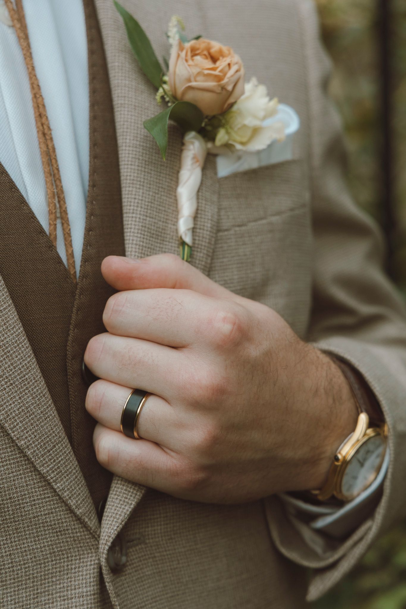 Close-up detail of a boutonniere with peach roses pinned to a tan suit jacket with wedding rings visible.