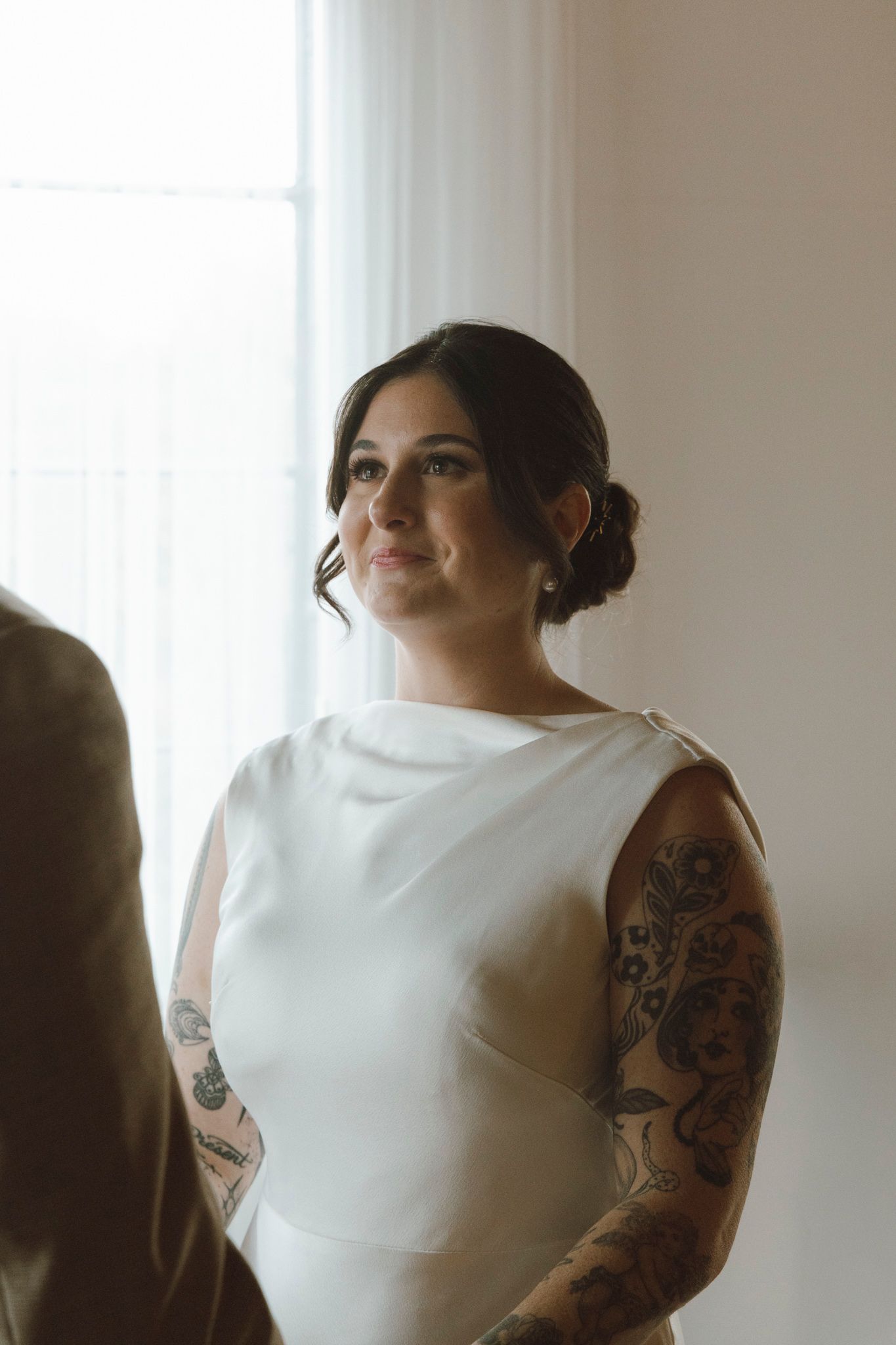 Person in white sleeveless wedding dress with visible arm tattoos poses near window in soft natural light.
