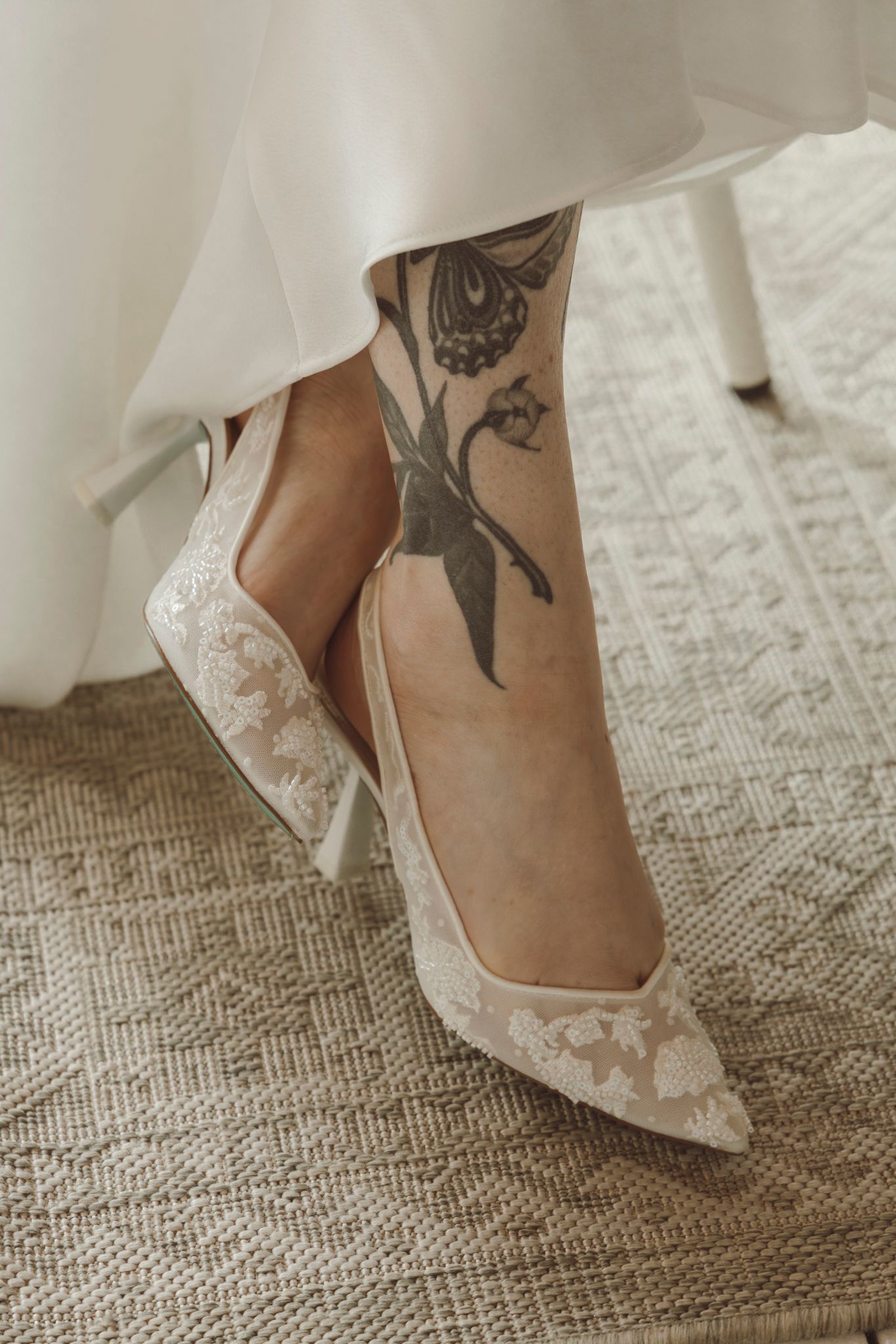 Lace wedding shoes with visible foot tattoos against textured carpet background.