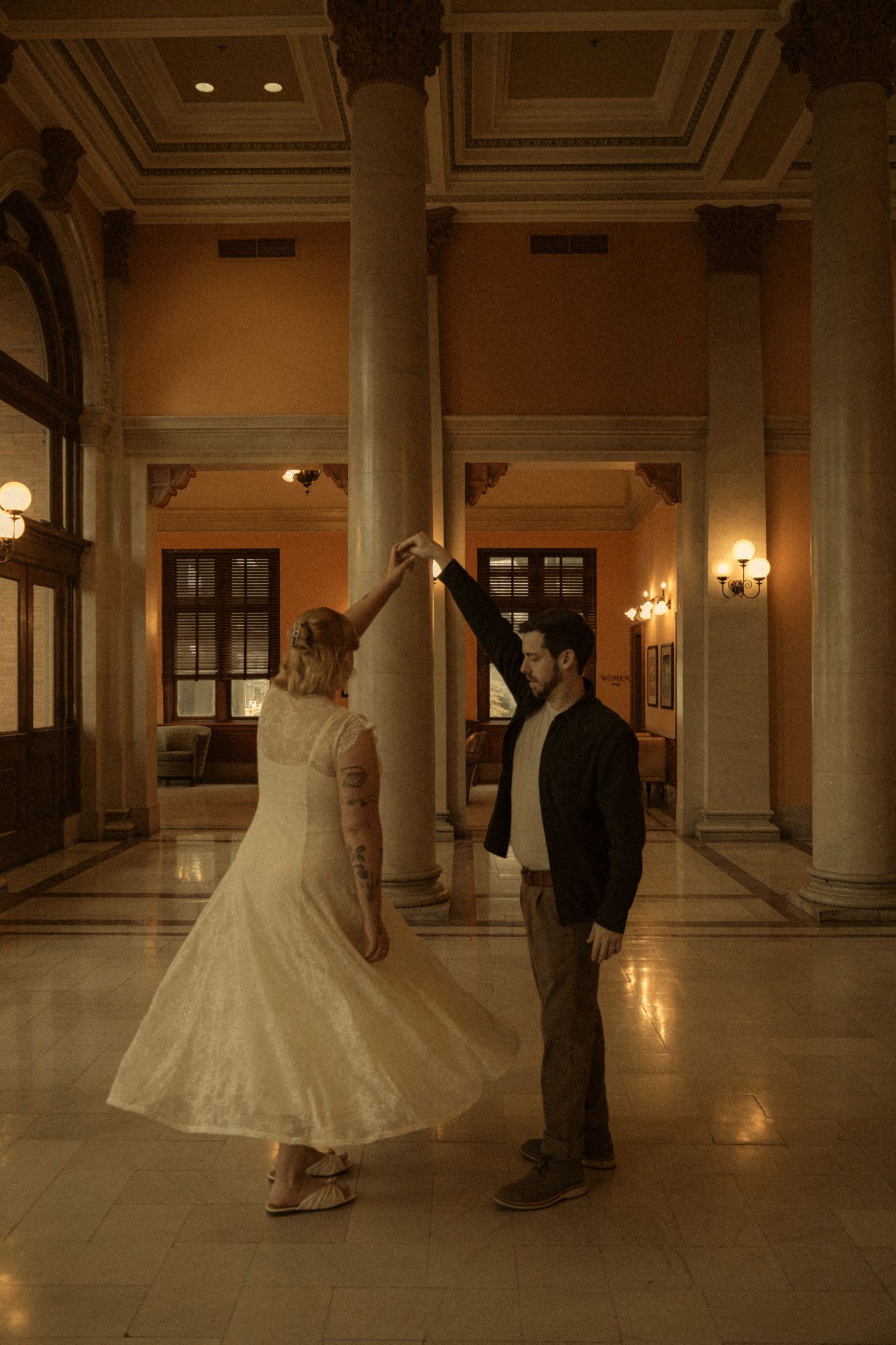 A couple in formal wedding attire dance together in an elegant, dimly lit hallway with ornate columns and architecture.