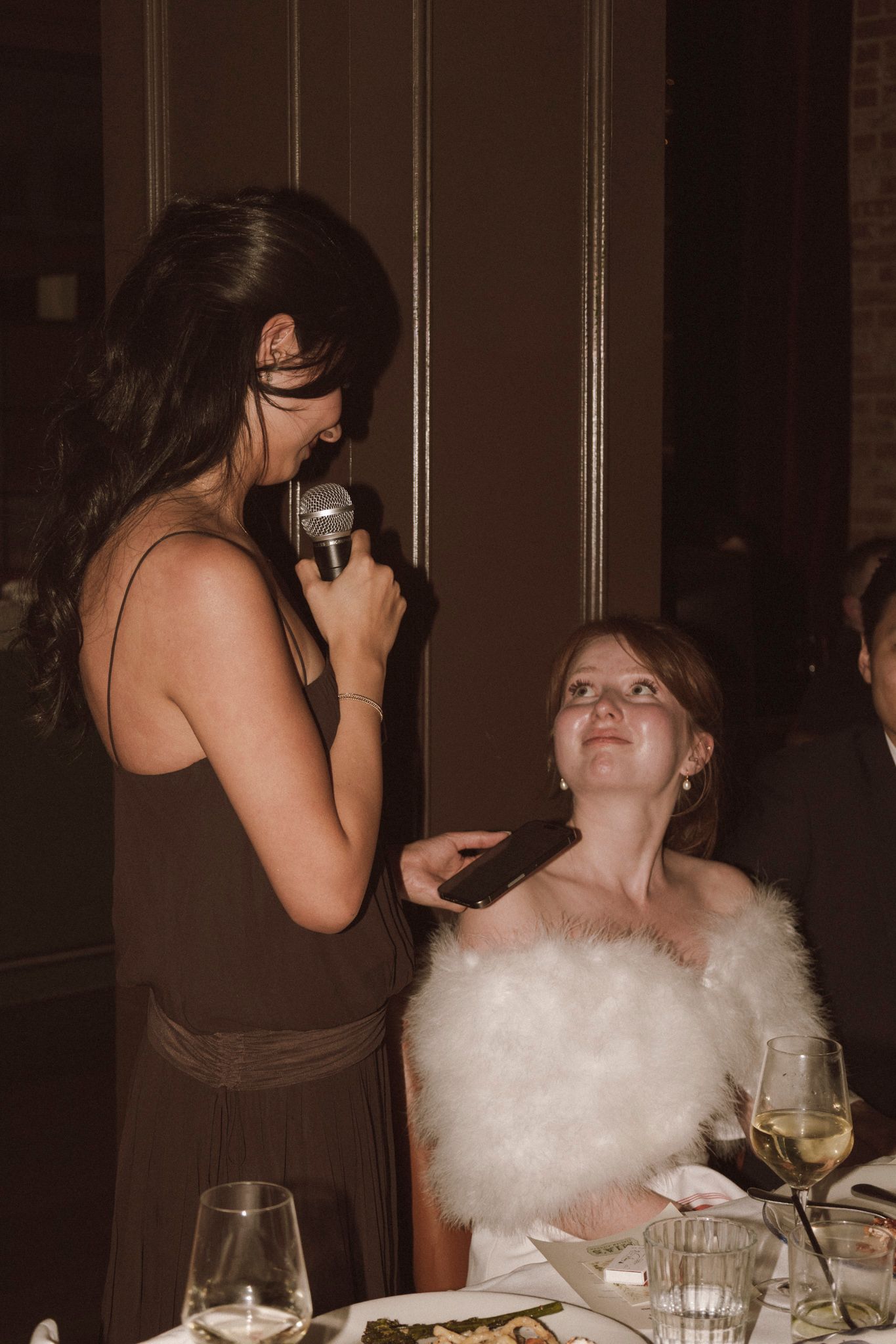 A bridesmaid in a dark dress holds a microphone while speaking to the bride at a wedding reception dinner.