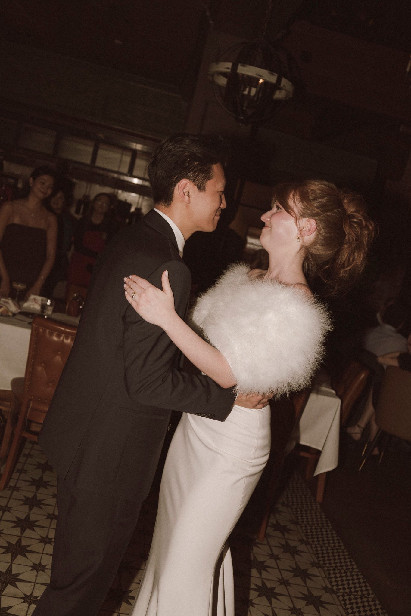 Bride in white gown with fur stole and groom in tuxedo share first dance at elegant wedding reception.