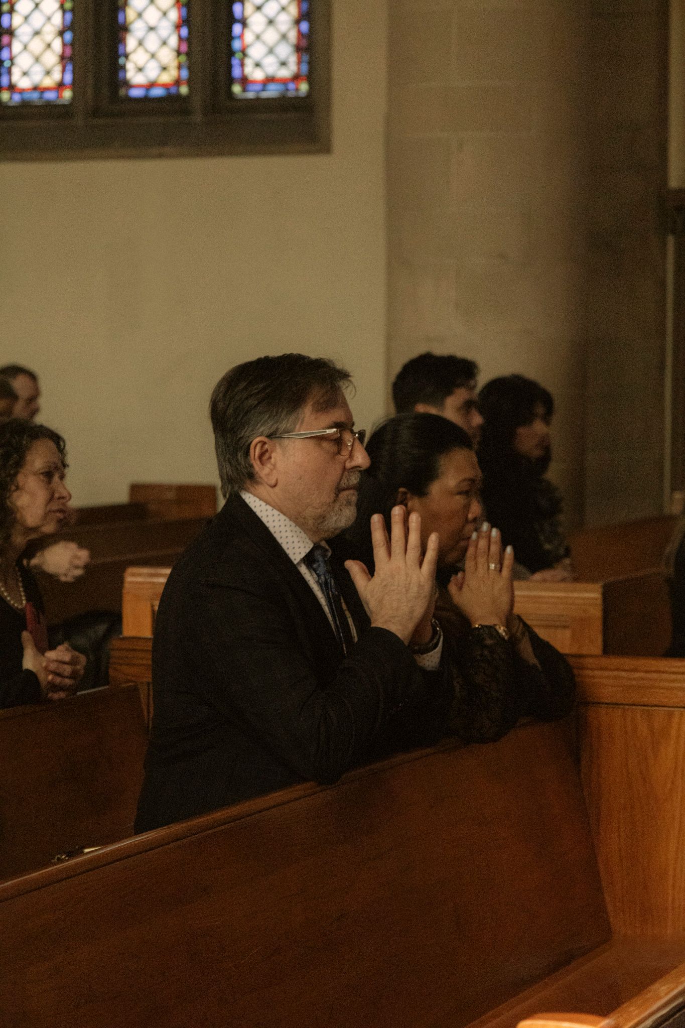 Man with hands clasped in prayer seated in church pew with other wedding guests during ceremony.