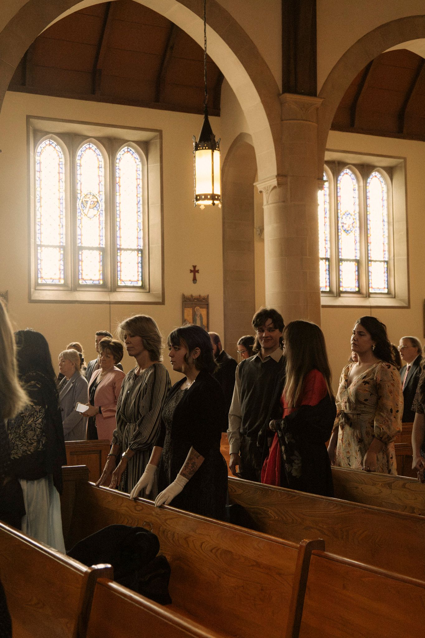 Guests standing in church pews during a wedding ceremony in a historic stone church with stained glass windows.