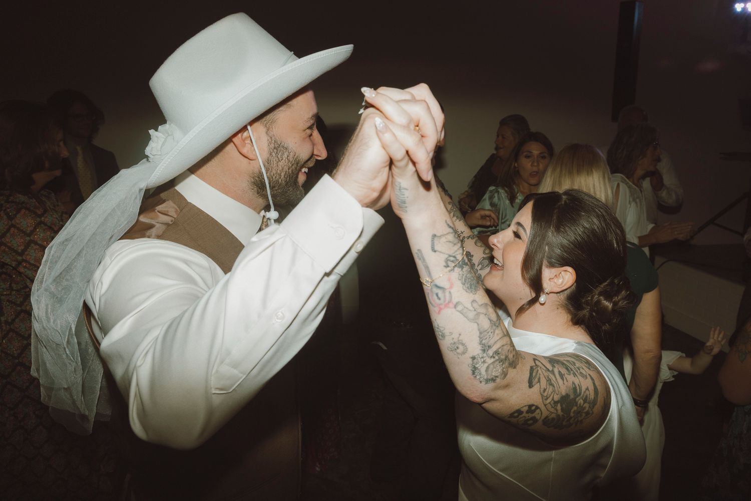 A couple dances together at a wedding reception, with one person wearing a white hat and the other showing tattooed arms.