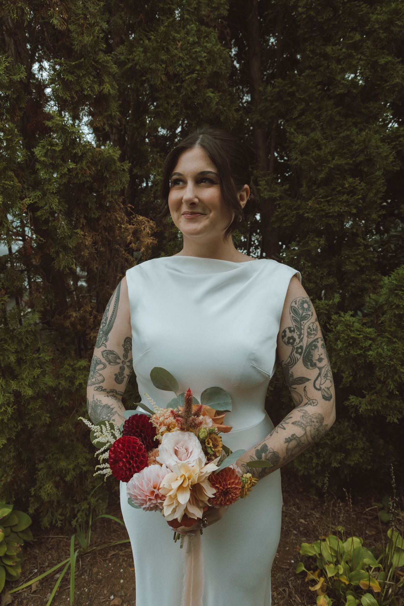 A bride in a sleek white dress holds a vibrant autumn bouquet with red and peach flowers against a wooded backdrop.