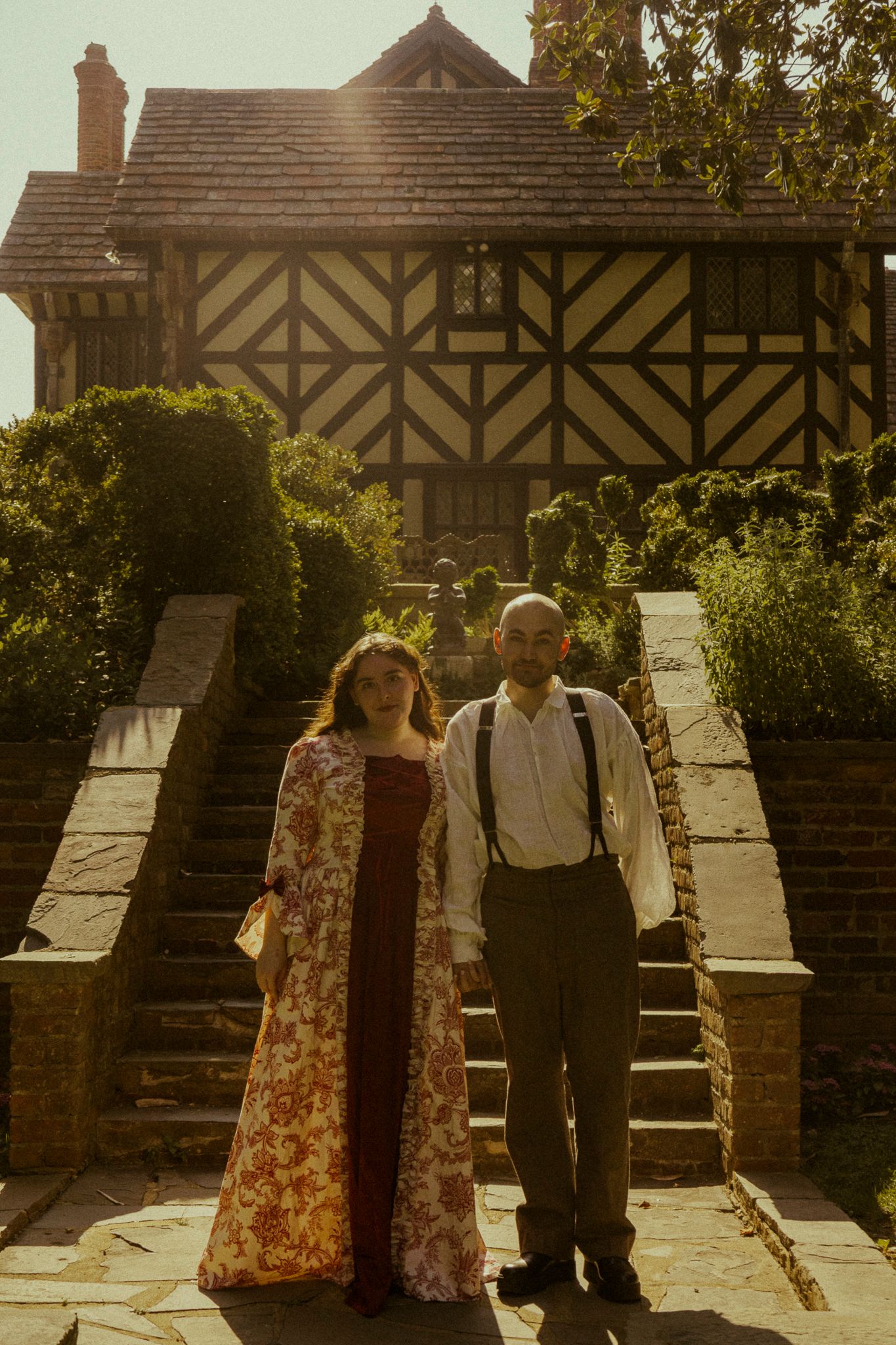Couple posing in front of a Tudor-style manor house with stone steps and lush gardens in golden sunlight.