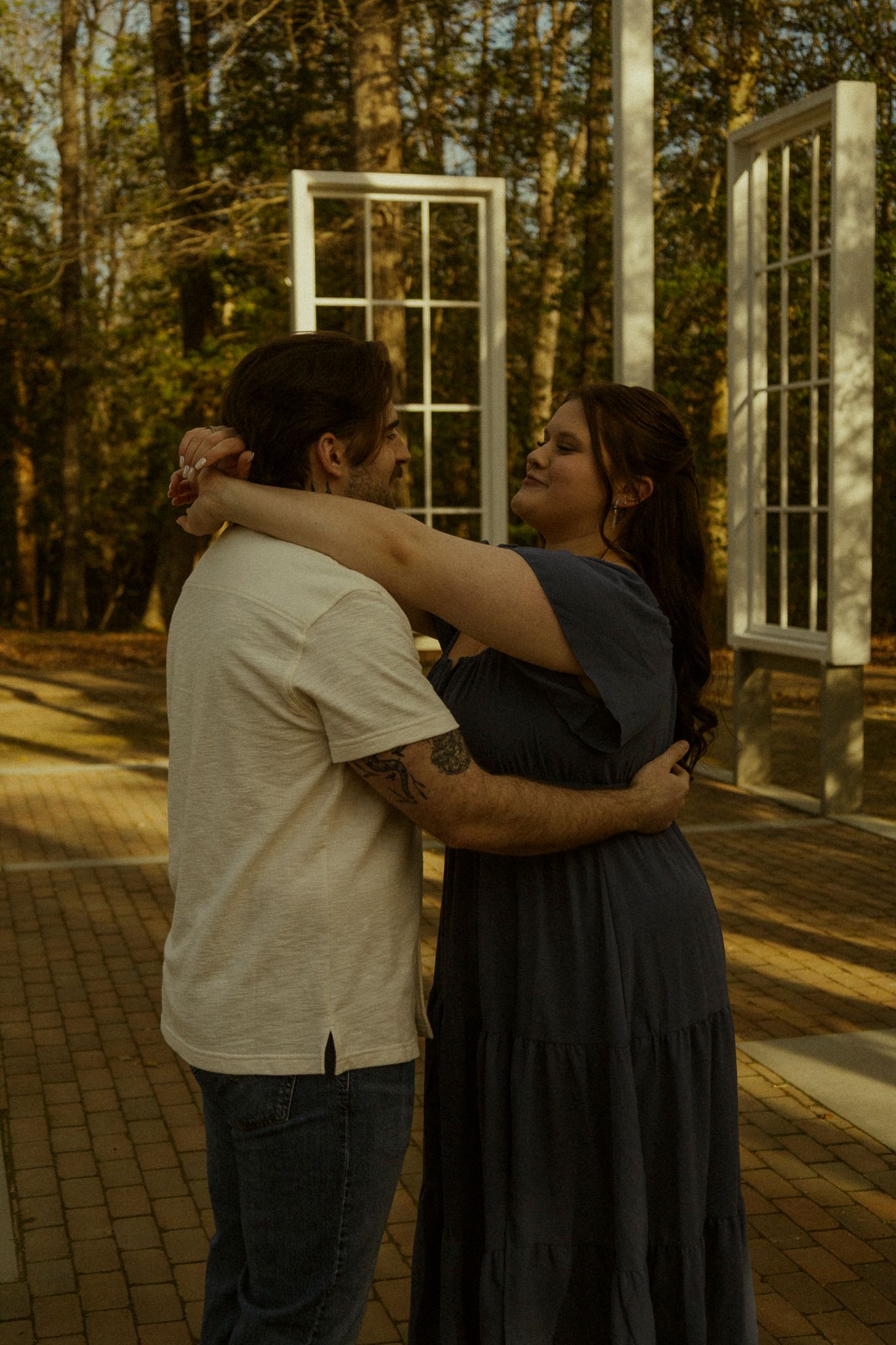 Couple embracing outdoors near white window frames among autumn trees on a brick pathway.