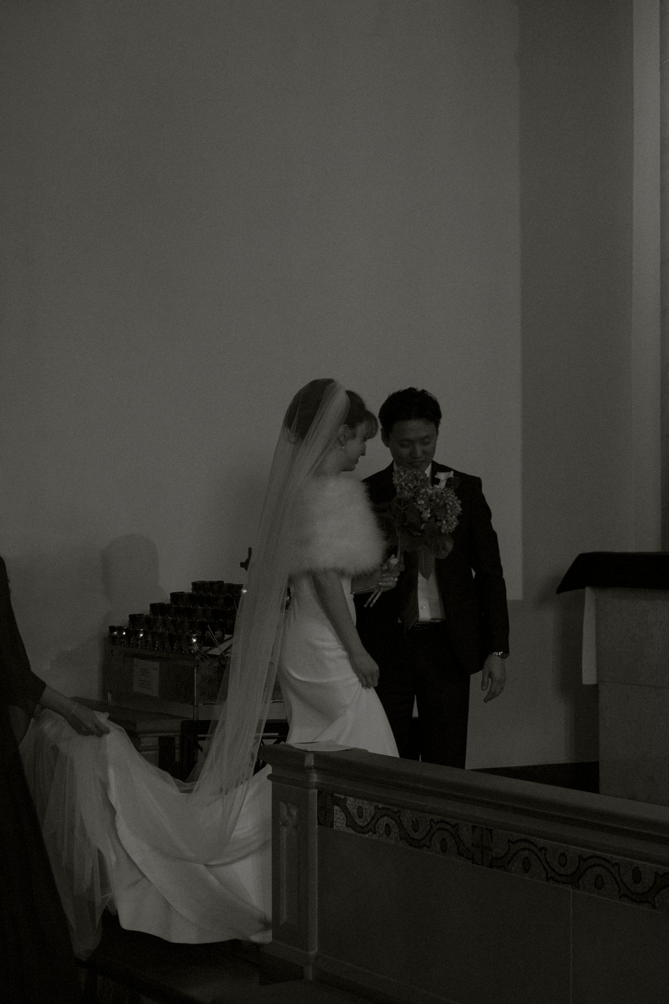 Black and white photo of bride in flowing veil and gown walking with groom holding bouquet along church pews.