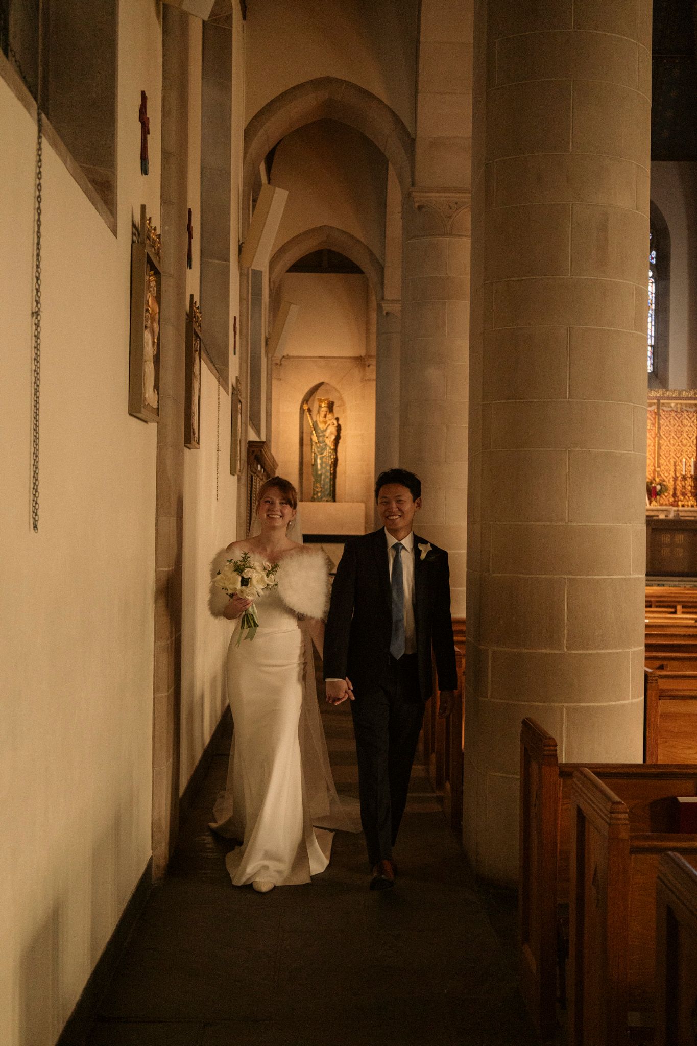 Newlyweds strolling together through a golden-lit historic stone church corridor after their wedding.