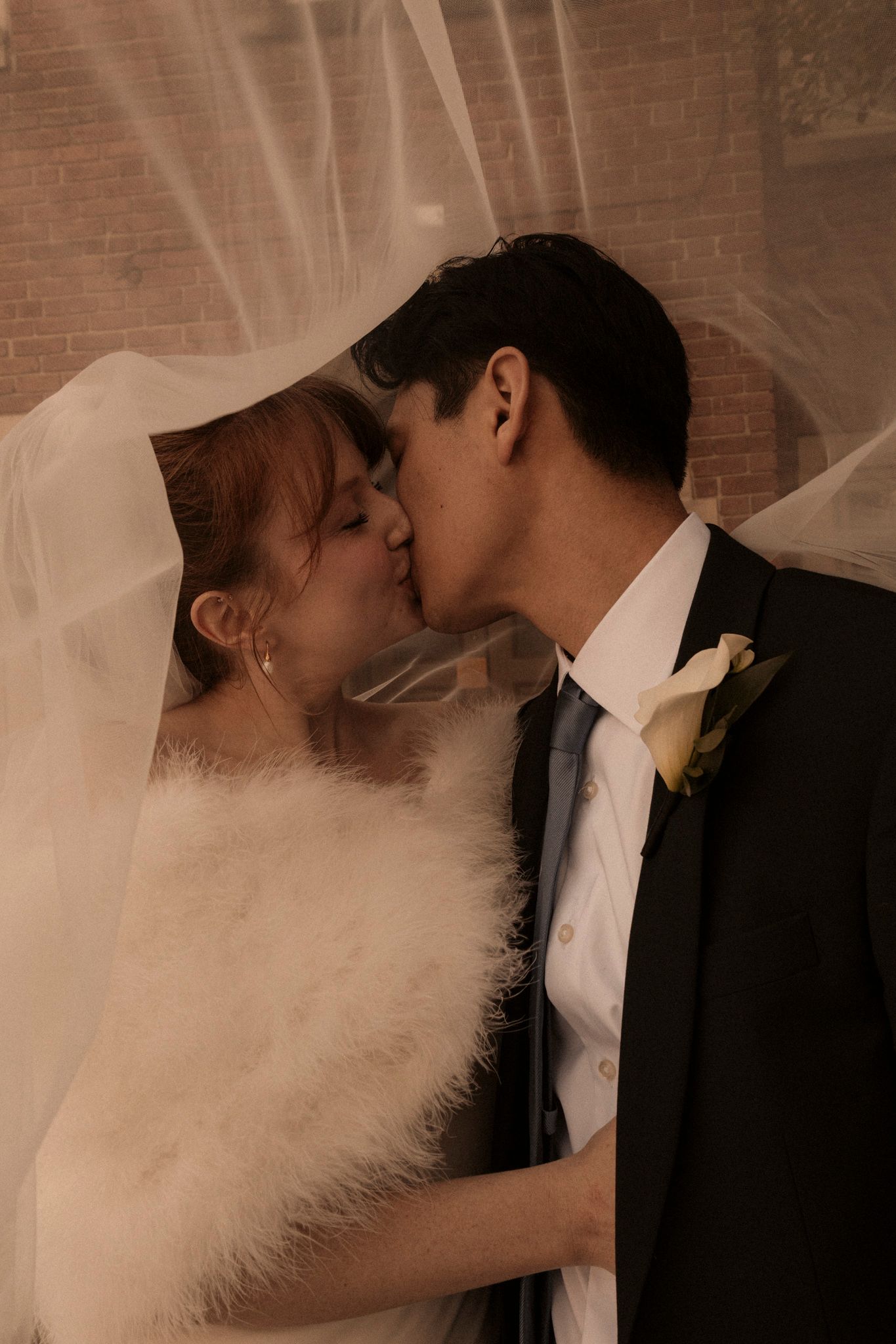 Bride and groom sharing a romantic kiss beneath a billowing veil, bride wearing a feather wrap.