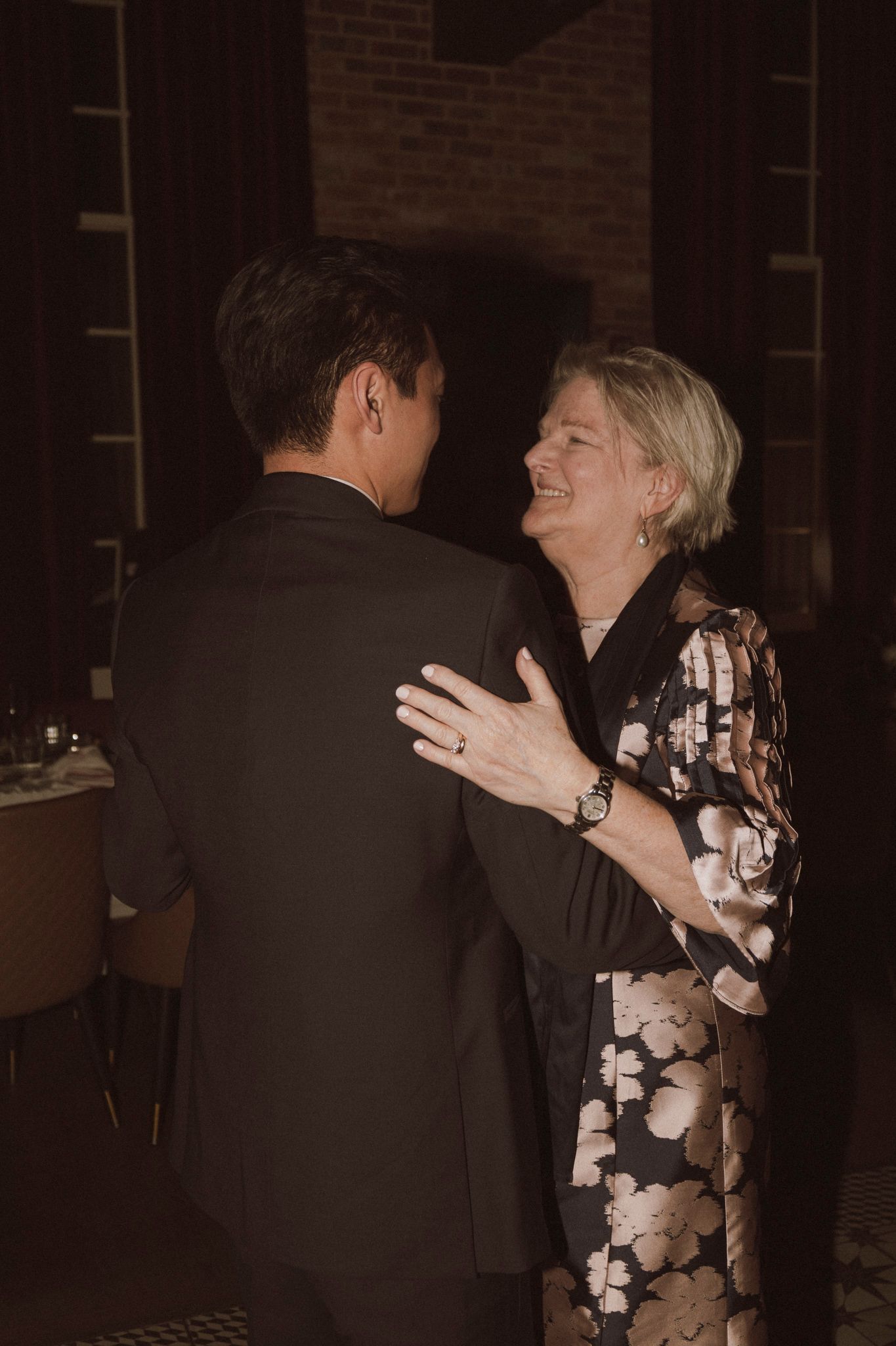 Groom dancing with older woman in floral dress at wedding reception, both smiling warmly.