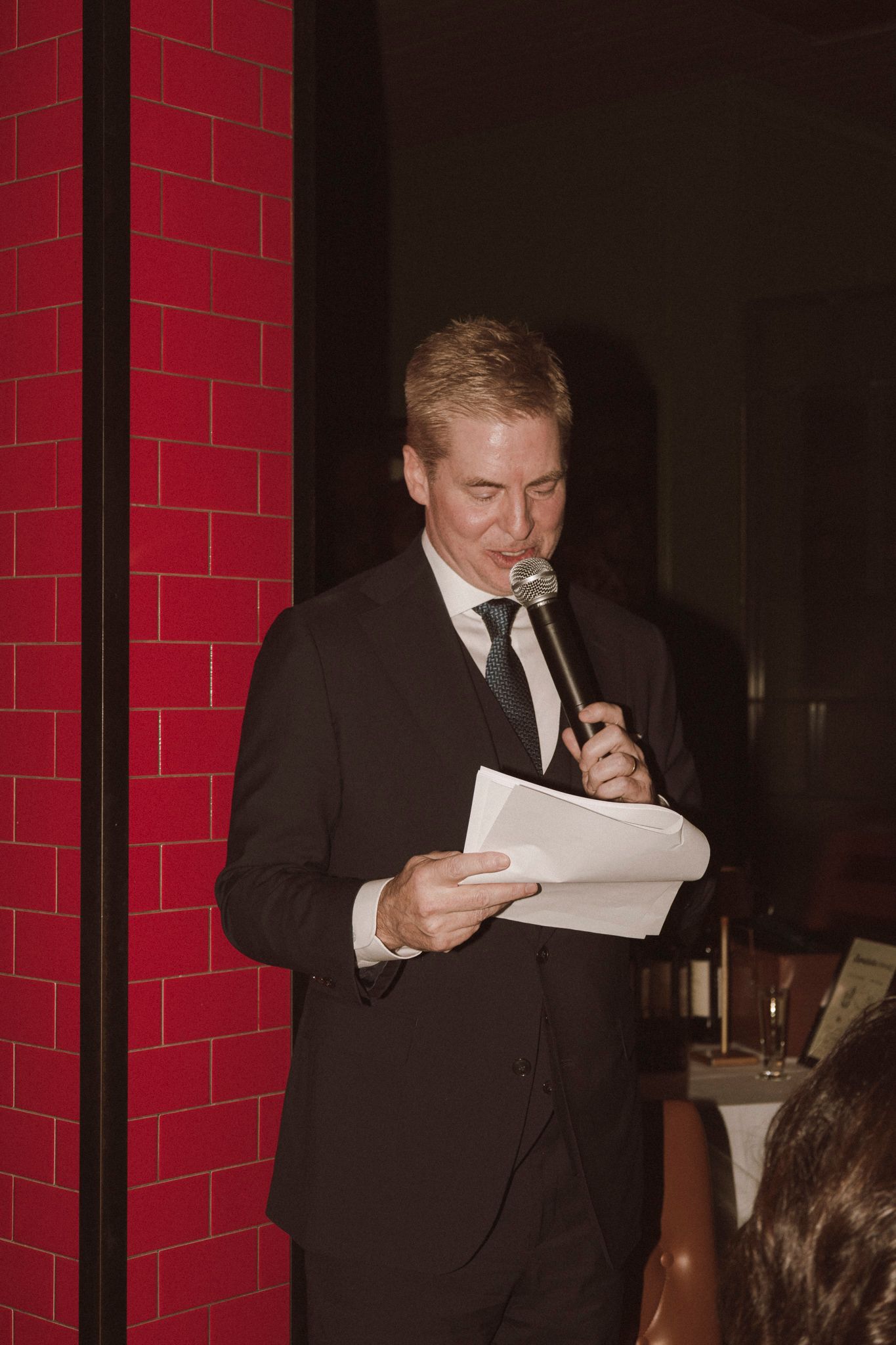 Blond man in black suit reading from papers while speaking into a microphone at a wedding reception.