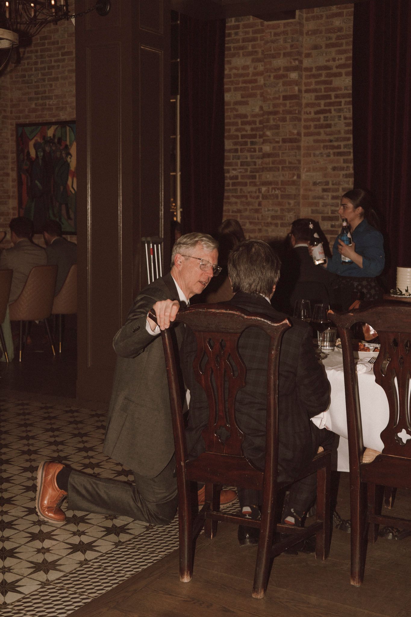 Elderly man kneeling on restaurant floor conversing with seated guest at formal dinner event.