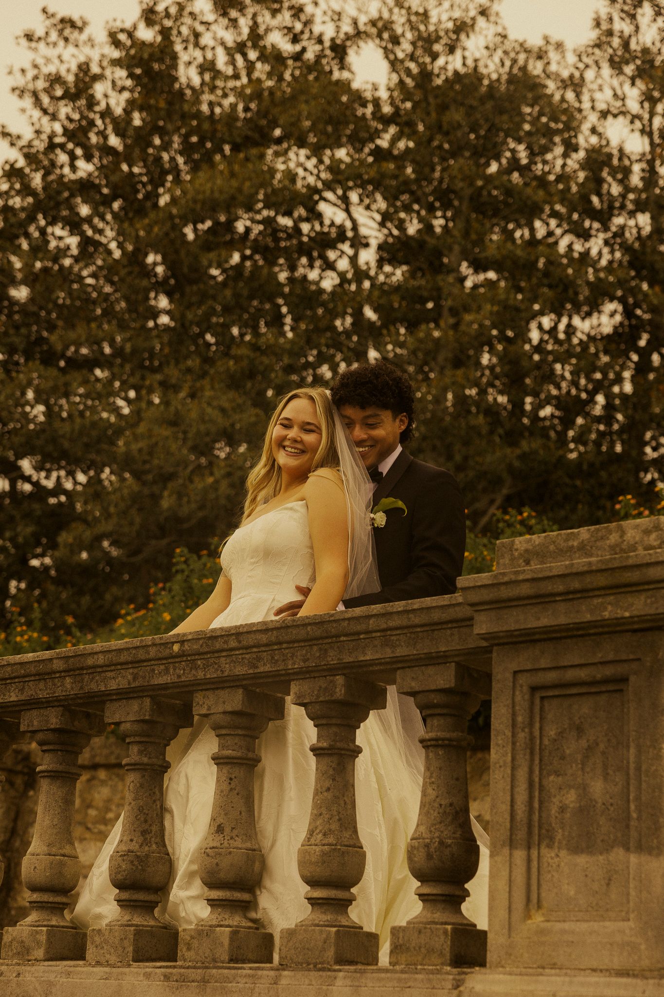 A smiling bride and groom pose together on a stone balustrade balcony surrounded by lush green trees.