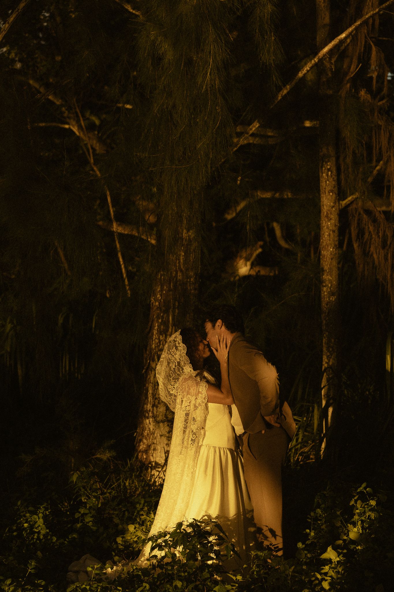 Couple sharing a romantic kiss in a dark forest at night, illuminated by warm golden light, bride in white gown.