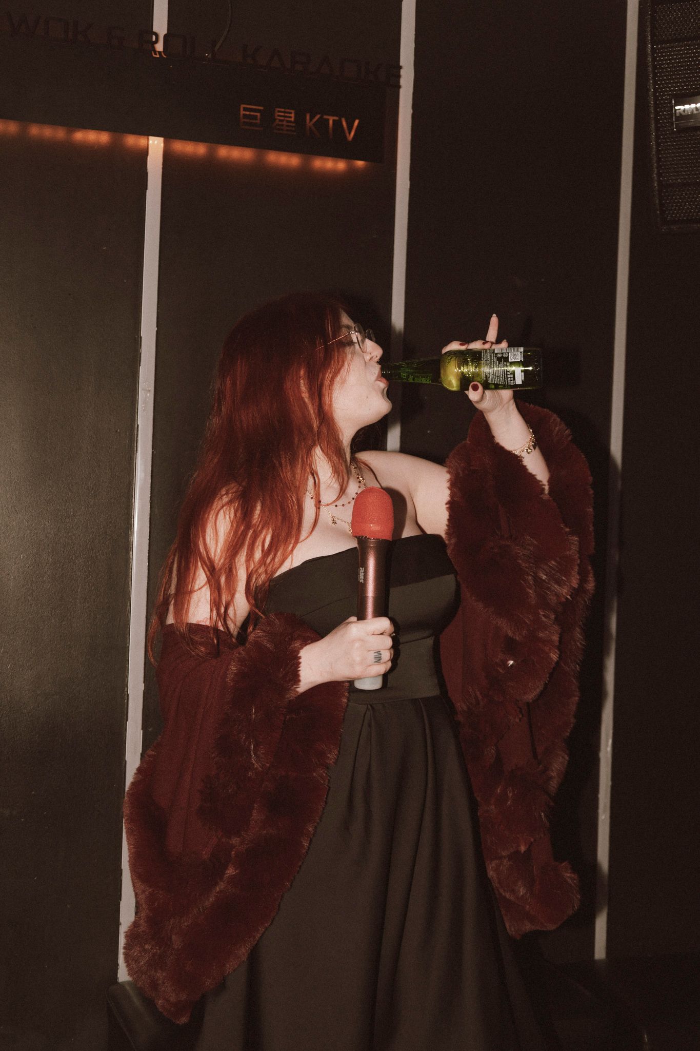 Person in a dark dress and burgundy fur coat holds a microphone and bottle in a dimly lit venue.