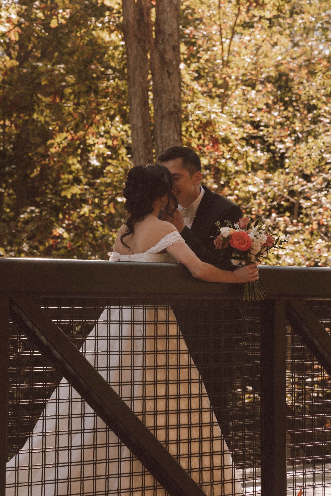 Newlyweds embrace on a rustic wooden deck with golden fall trees creating a romantic backdrop.