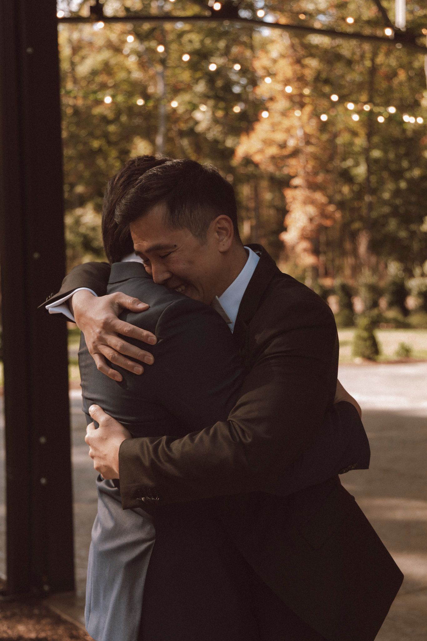 Two people in formal black attire share an emotional embrace outdoors during a special moment.