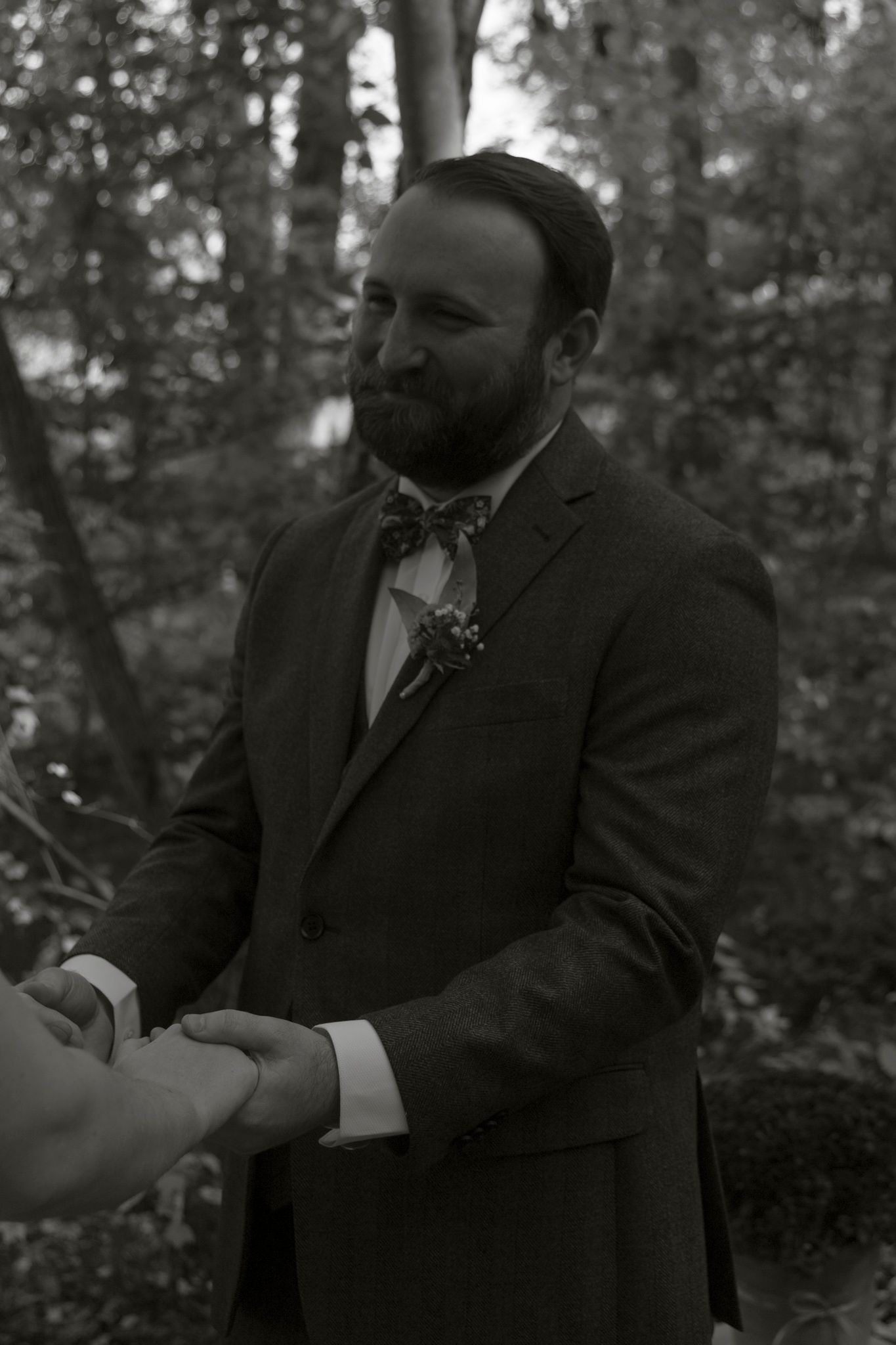 Black and white photo of a bearded person in a suit and bowtie shaking hands outdoors in a wooded setting.