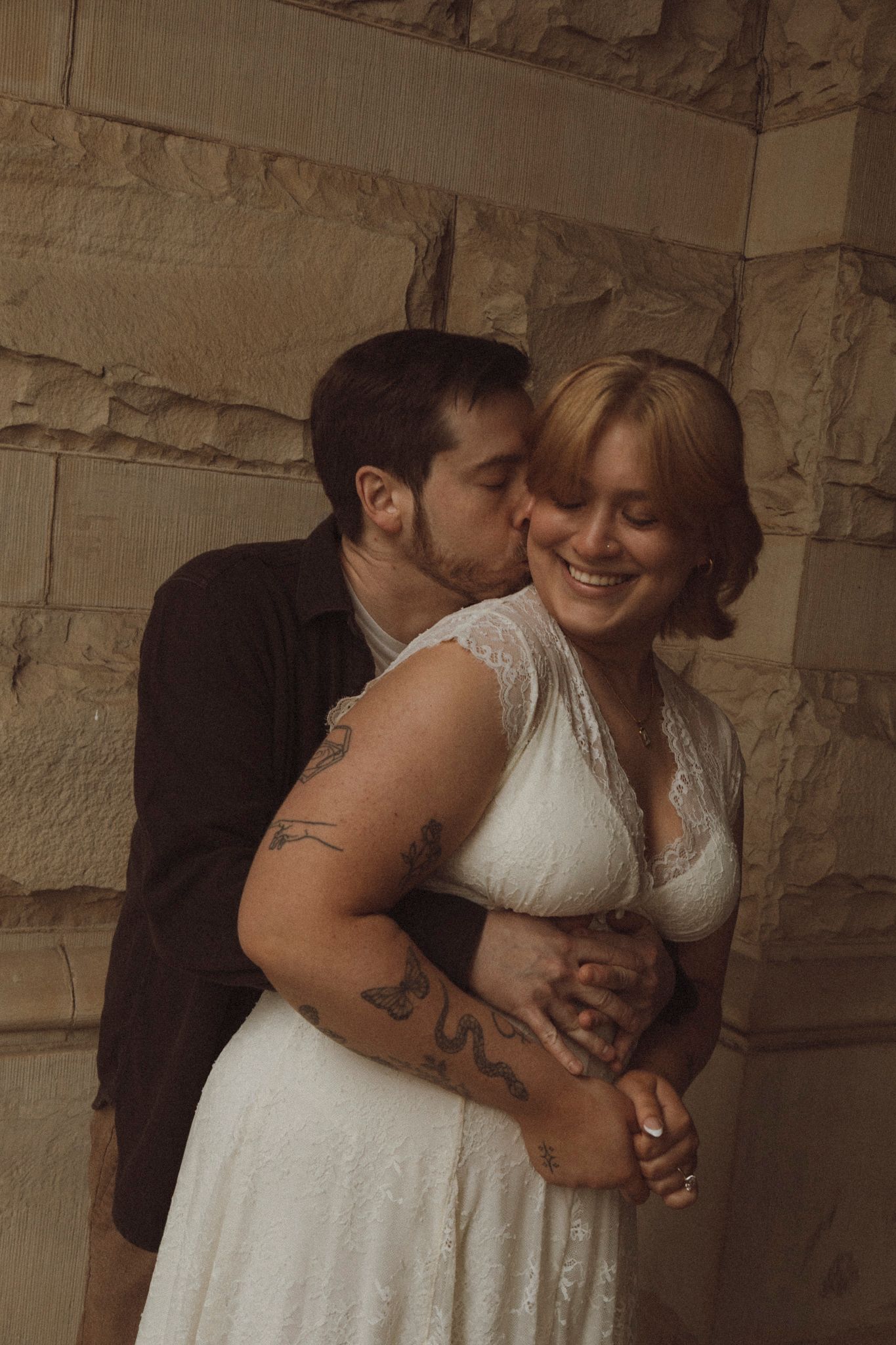 A romantic moment between newlyweds embracing against a stone wall, with the bride wearing a white lace dress.