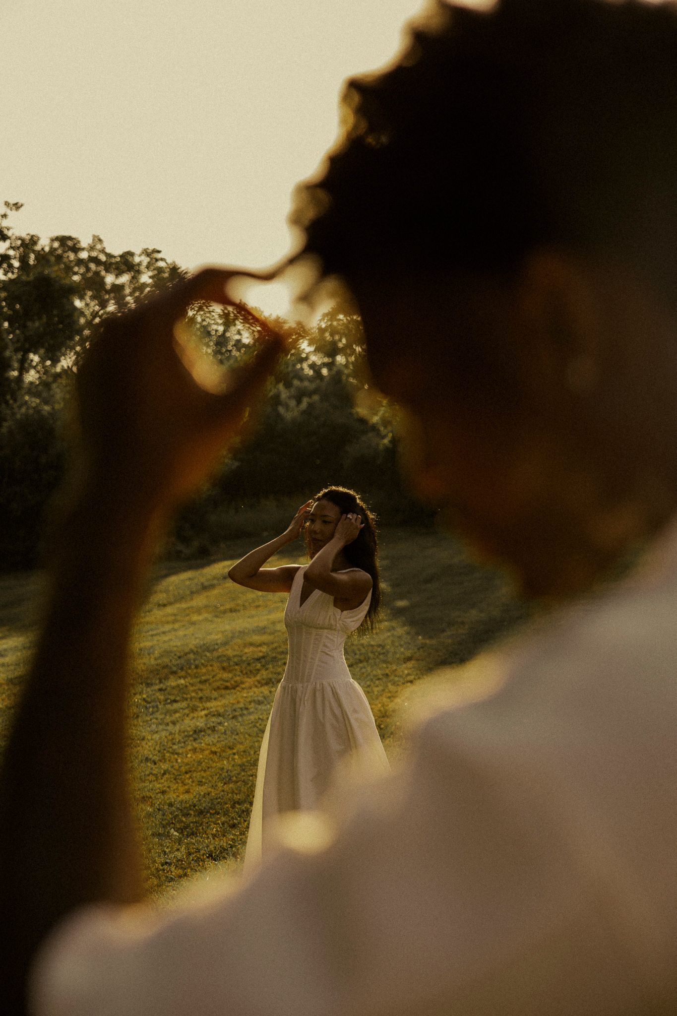 Man forming heart shape with hands framing woman in white dress standing on sunlit green hillside.