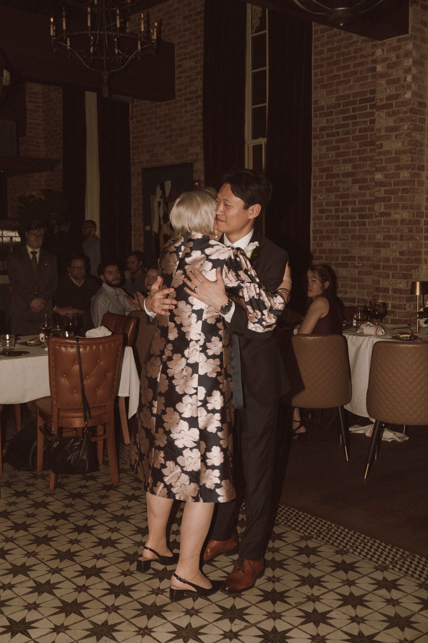 Groom in dark suit shares a dance with older woman in floral dress at wedding reception.