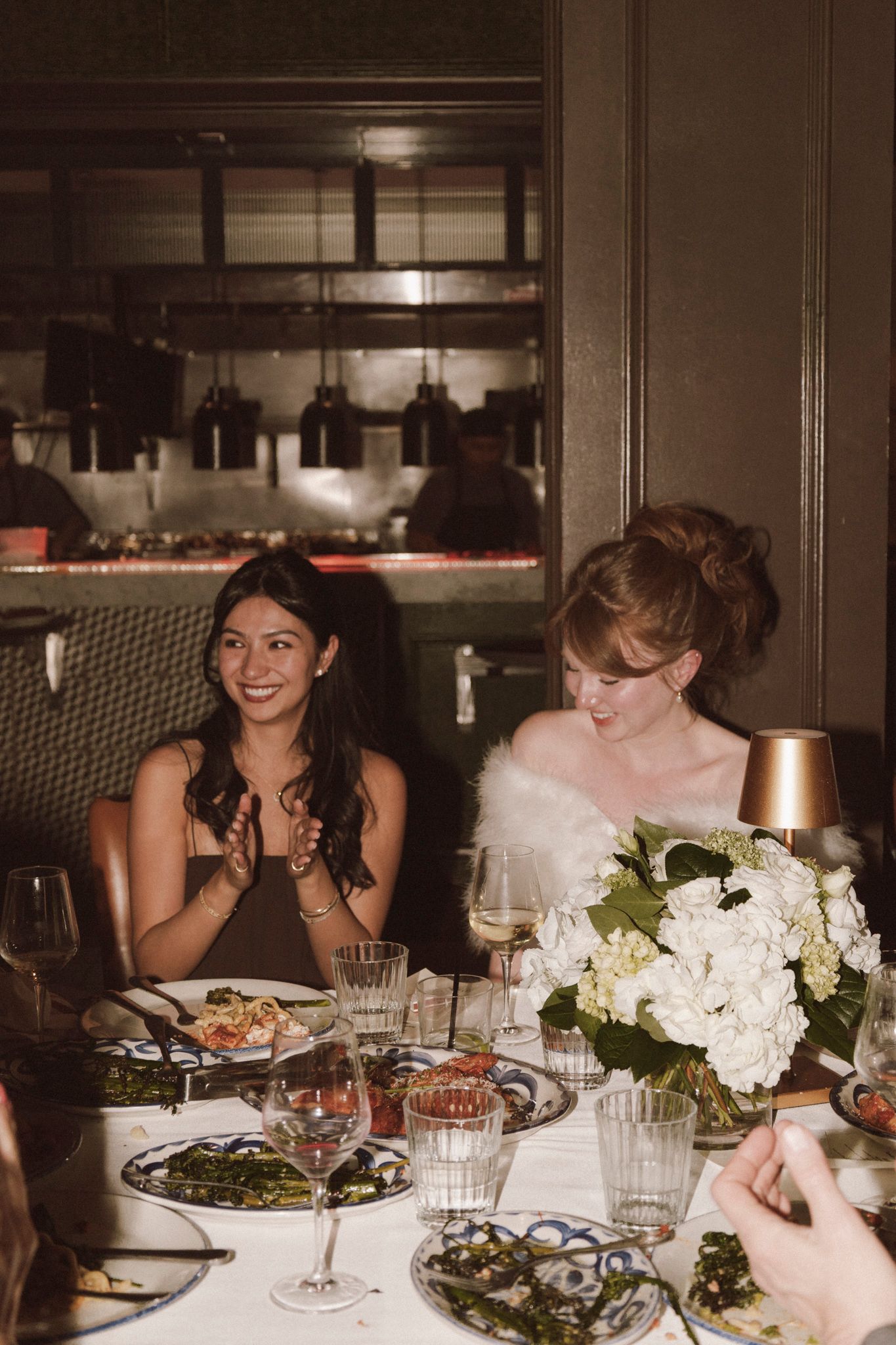 Two women at wedding reception dinner, one clapping beside bride with white floral bouquet.