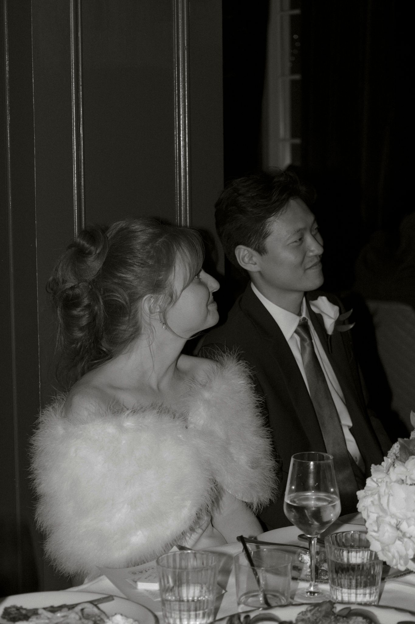Black and white portrait of a bride in a white fur stole and groom in a tuxedo at their wedding reception.