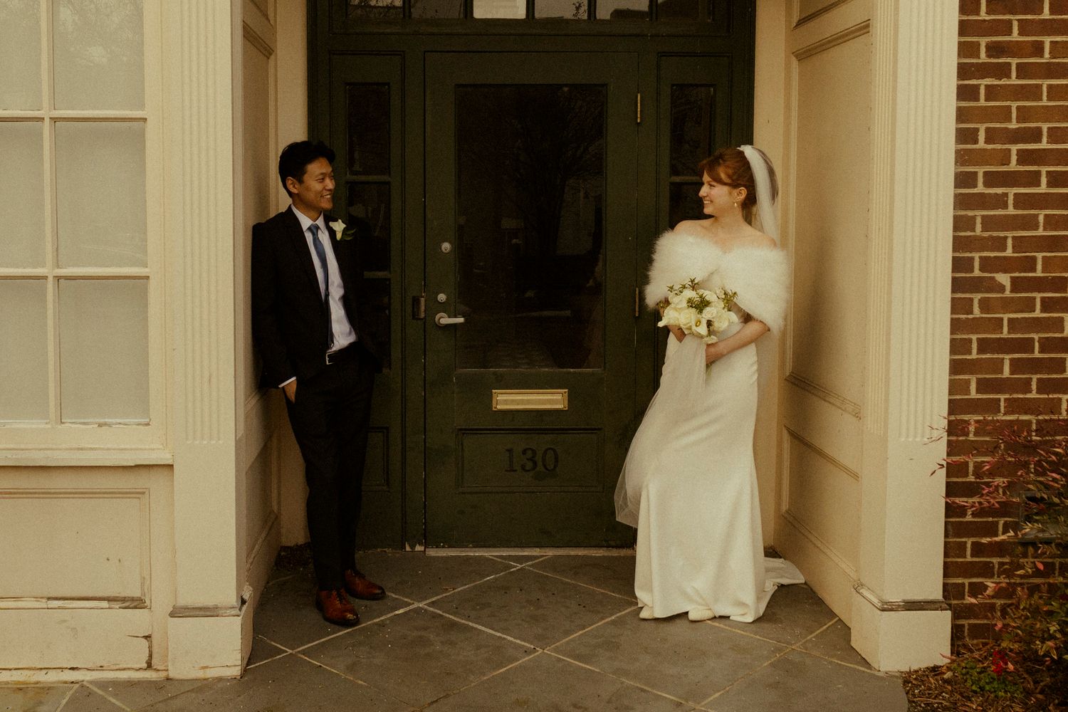 Bride holding bouquet and groom share a first look moment outside an elegant brick building.
