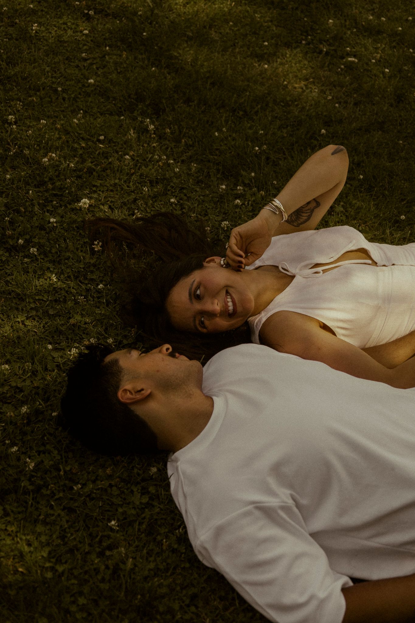 Couple lying on green grass smiling at each other, woman in white dress with tattoo, man in white shirt.